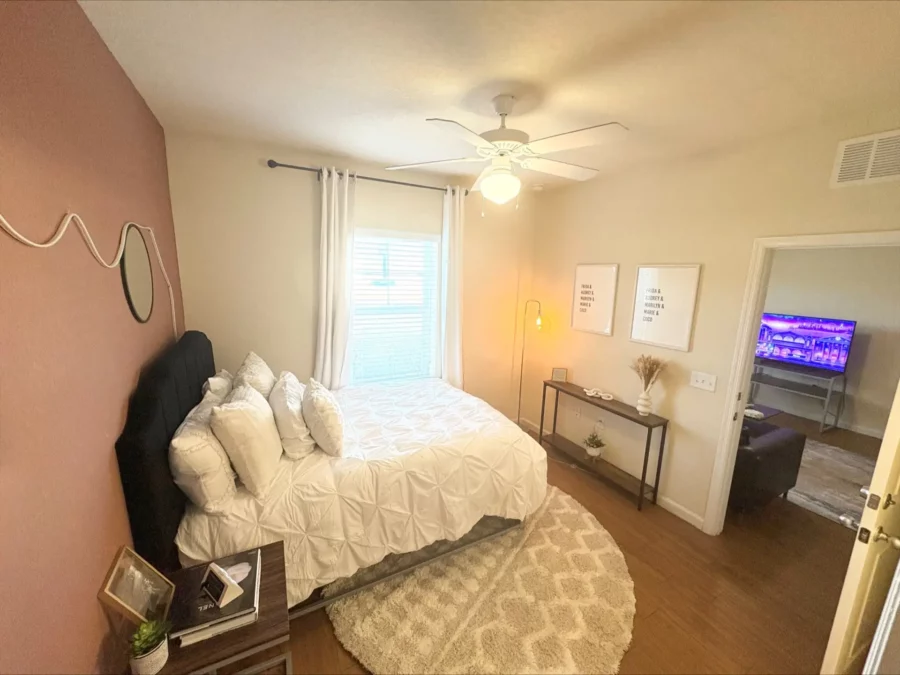 apartment bedroom with wood style flooring, a tall bed with storage underneath and side table, a narrow table against the wall, and large window.